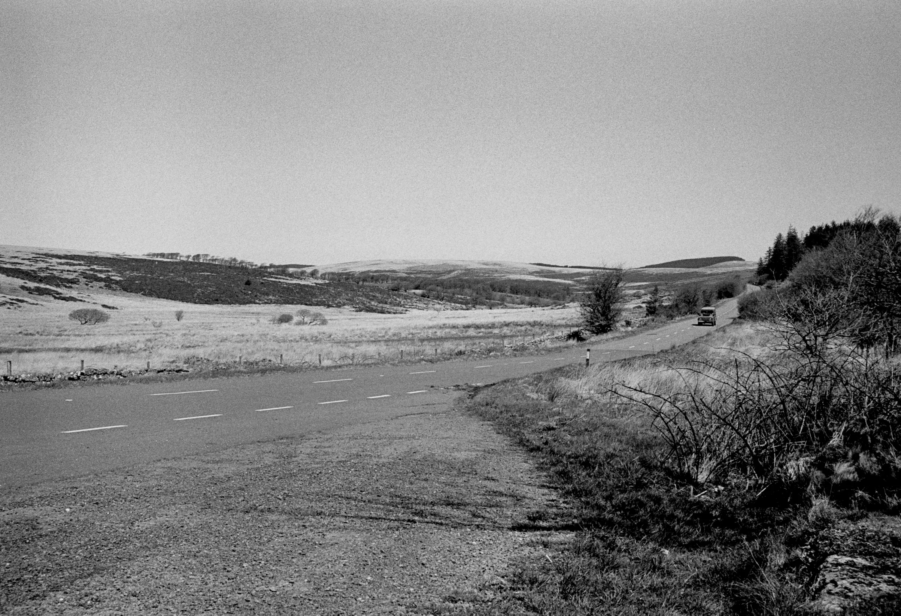 A road curving away across open moorland in black and white, a single car in the distance, rough scrub in the foreground and low hills on the skyline.
